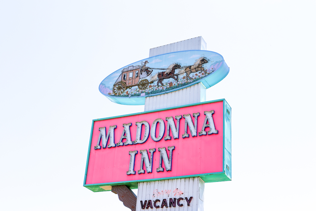 Roadside neon sign for the Madonna inn with a image of stagecoach and horses on blue background above the neon letter spelling Madonna Inn. Sorry no vacancy is lit up in neon.