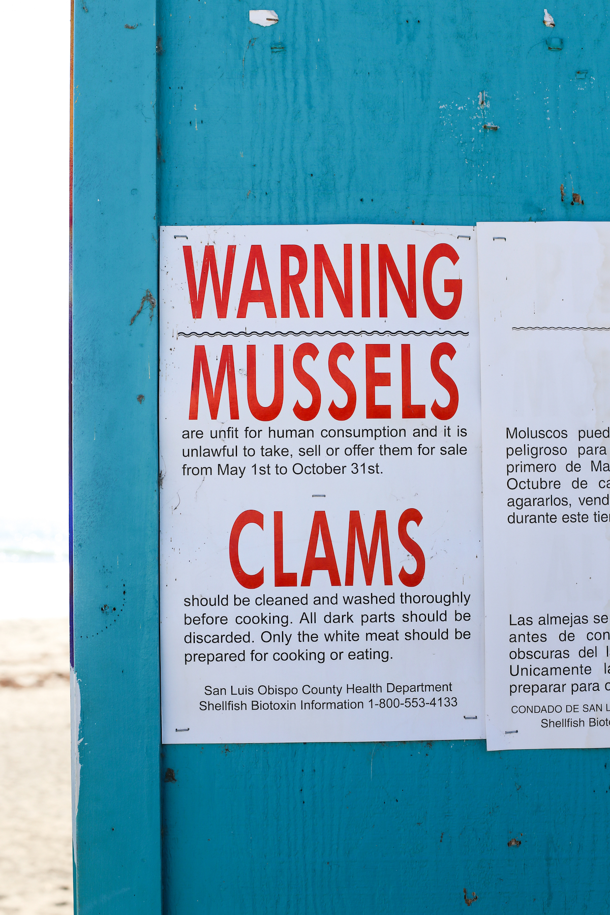 Sign posted on blue building reads "Warning: Mussels are unfit for human consumption and it is unlawful to take, sell, or offer them for sale from May 1st to October 31st. Clams should be cleaned and washed thoroughly before cooking. All dark parts should be discarded. Only the white meat should be prepared for cooking or eating. 
San Luis Obispo County Health Department Shellfish Biotoxin Information 1-800-553-4133